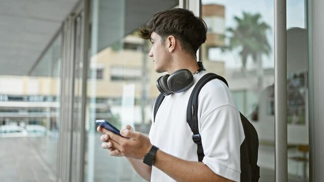 Young hispanic teenager student wearing headphones and backpack using smartphone at university