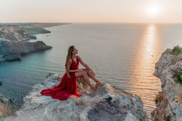 Woman sunset sea red dress, side view a happy beautiful sensual woman in a red long dress posing on a rock high above the sea on sunset. - obrazy, fototapety, plakaty