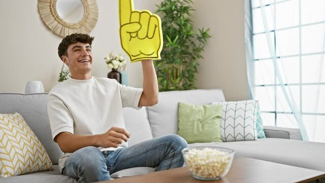 Excited young hispanic male teenager, avid soccer team fan, joyfully watching the match on tv, comfortably sitting on living room sofa at home, savouring tasty popcorn snack.