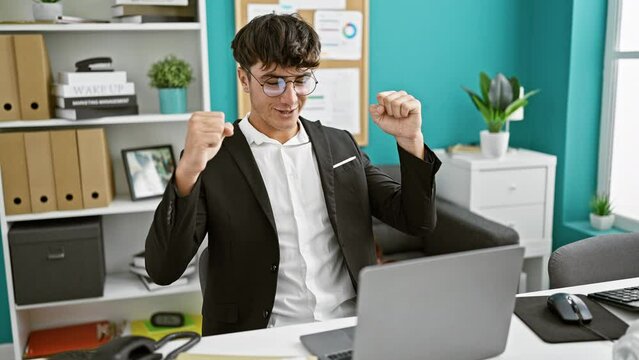 Young hispanic teenager celebrating business success at the office, brimming with confidence, crossing fingers for luck on laptop work