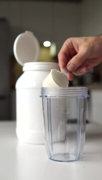 Young man with measuring spoon in his hand puts portion of whey protein powder into a shaker on wooden table, making protein drink. Vertical video.