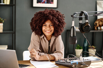 Happy young African American female host looking at camera while sitting by workplace with...