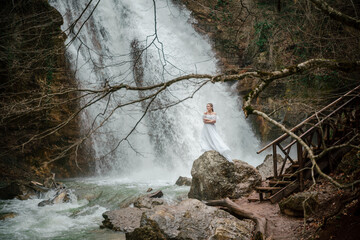 Happy woman in a white dress stands on a stone with a waterfall behind