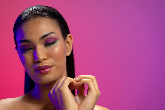 Biracial woman with dark hair, heart of hands, pink lips, eyeshadow make up on neon background