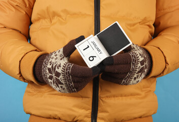 Man in yellow winter down jacket and gloves holding block calendar with date january 16 on blue...