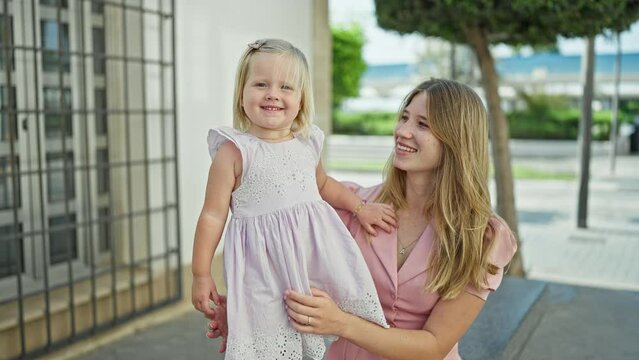 Joyful caucasian mum confidently standing on street, arms cradling her giggling daughter, both sharing a fun, positive outdoor moment in their favorite city.