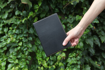 Black leather notebook in woman’s hand against the green leaves wall background