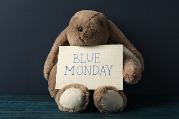 Plush bunny with paper with text Blue Monday on wooden table on dark blue background, close up