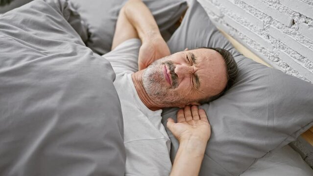 Serious unhurt middle age man, with grey hair and glasses, distressed by cervical pain, lays awake, touching neck in bed - a heartbreaking morning sight in the background of his bedroom.