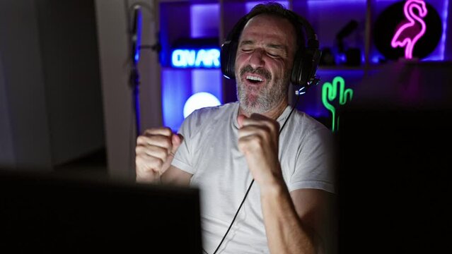 Cheerful middle age man with grey hair ecstatically celebrates his win on a video game stream in a dark gaming room