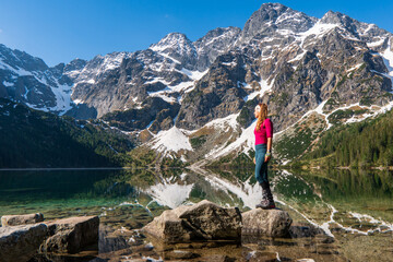 Young woman in red sportswear standing in the nature, mountain lake and range on the background, active life concept, summer time in Eye of the Sea lake in Tatra Mountains in Poland - obrazy, fototapety, plakaty
