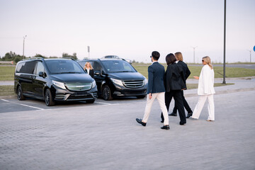 Group of an elegant business people walk together to minivan taxies on a parking lot. Concept of...