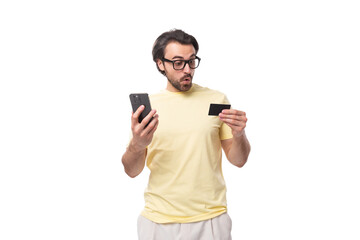 casual caucasian guy with a beard in glasses dressed in a light t-shirt holds a phone and a credit card