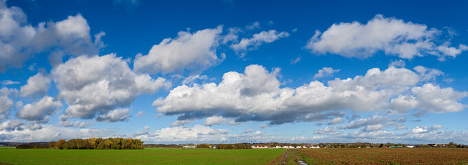 Panoramafoto von Cumulus- oder Haufenwolken am blauen Himmel in herbstlicher Jahreszeit mit Feldern, einer Siedlung und einem Wald am Horizont