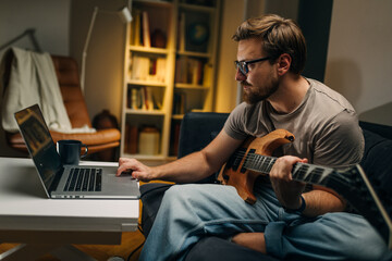 Side view of a Caucasian man with an electric guitar looking at notes on laptop - obrazy, fototapety, plakaty