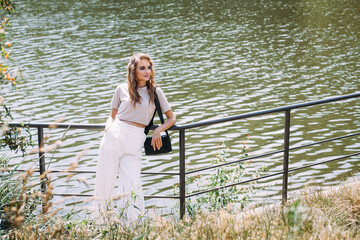 A beautiful girl with fashionable make-up and hairstyle, in white trousers and a beige top, with a black leather handbag, stands by the iron fence near the river on a hot sunny day.