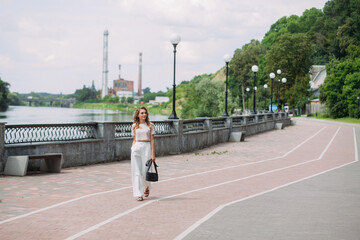 A slender young girl with fashionable make-up and hairstyle, in white trousers and a beige top and a black leather handbag, walks along the city embankment near a picturesque river.