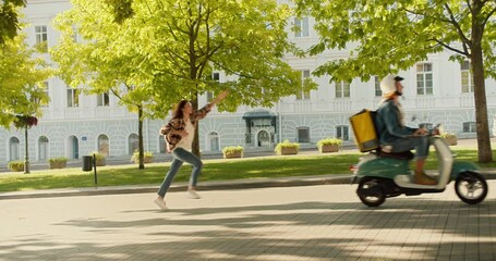 A funny situation: a brunette girl in a checkered shirt is running after a food delivery guy who is leaving her on a moped and does not hear her as she runs after him. The food delivery guy leaves his