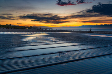 Beautiful dark golden sunset at golden hour at the beach next to dark and cold sea at summer right after the storm and rain reflected in puddle on wooden platform panels - obrazy, fototapety, plakaty