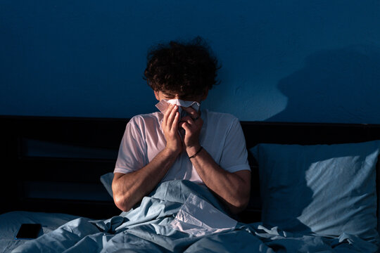 Muslim man suffering cold and flu trying to sleep. Young sick ill arabic guy with runny nose in casual clothes sitting in bed