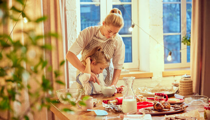 Beautiful, caring mother cooking with her little daughter at home in kitchen in evening. Teaching child to bake cookies. Concept of cookie day, motherhood, childhood, holidays, family - obrazy, fototapety, plakaty