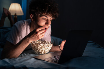 Happy muslim man eating popcorn while lying on bed watching movie on laptop at night at home.