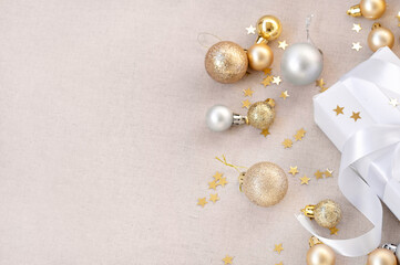 White gift box fragment, golden and silver pastel Christmas decoration balls, ornaments, sparkling garland lights and star confetti on light neutral beige or pink linen cloth