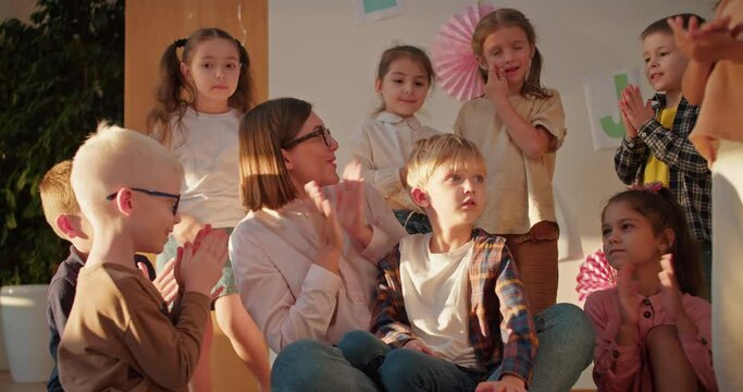 a female teacher with a bob hairstyle with glasses in a white shirt claps for a little blond boy along with the rest of the group of children in the pre-school club