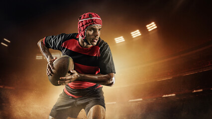 Concentrated young man, rugby player in uniform with ball standing on dark empty field with flashlights and mist. Concept of professional sport, competition, motivation, game, championship