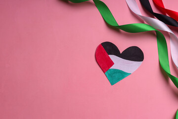 A heart-shaped paper and ribbons with colors representing the Palestinian flag over pink background
