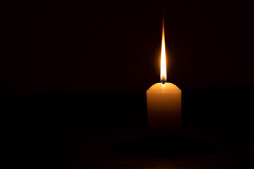A single burning candle flame or light glowing on a yellow candle isolated on red or dark background on table in church for Christmas, funeral or memorial service - obrazy, fototapety, plakaty