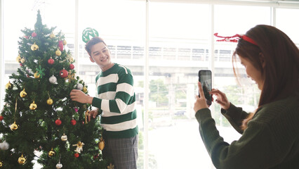 Happy young joyful Asian couple having fun taking photos, decorating various baubles on the...