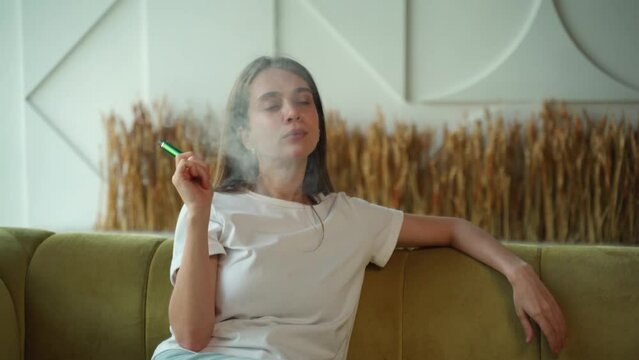 Beautiful young woman using disposable electronic cigarette on sofa at home. 