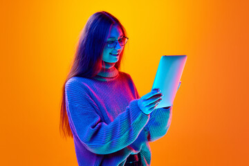 Portrait of cheerful girl, student dressed wool sweater holding tablet and chatting online in neon light against gradient background.