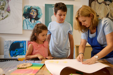 Charming female painter artist, instructor teaching assisting elementary age kids in painting at...