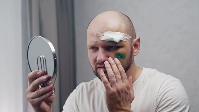 A wounded man with a stitched incision and a medical bandage on his forehead looks in the mirror, touches the wounds and sobs in pain. Concept consequences of a car accident
