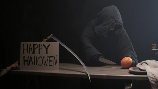 Grim Reaper sitting at a table with a Happy Halloween sign and ominously cuts a pumpkin with a knife. Halloween theme.