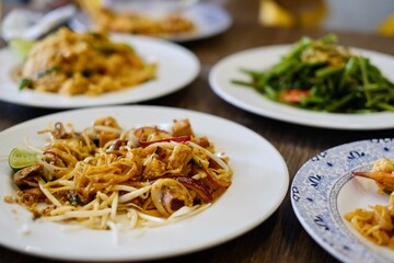 A full table of Thai dishes: seafood pad thai, chicken tom yum fried rice and stir-fried morning glory at a beachfront restaurant in Patong Beach, Phuket - Thailand