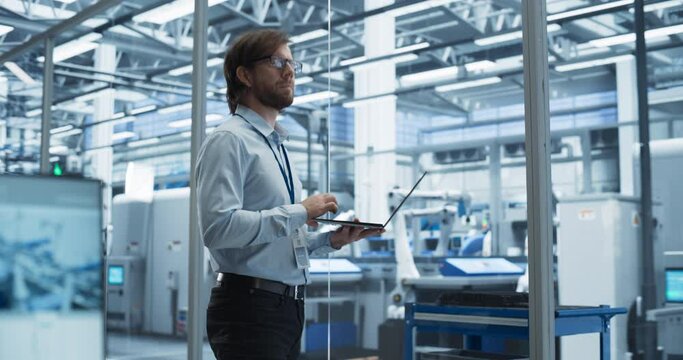 Portrait of a Young Factory Manager Working on Laptop at an Industrial Manufacture with Autonomous Assembly Robots. Specialist Monitoring Production on a Computer, Planning Workload for the Shift
