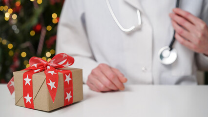 Medical banner concept for Christmas and New Year.Female doctor in white coat sits at her desk,on table there is gift box with red bow against background of beautifully decorated Christmas tree.