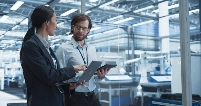 Portrait of a Cheerful Engineer and Happy Asian Scientist Using Laptop and Tablet Computers, Talking in a Factory Facility with Robots Producing Modern Electronic Components for Different Industries