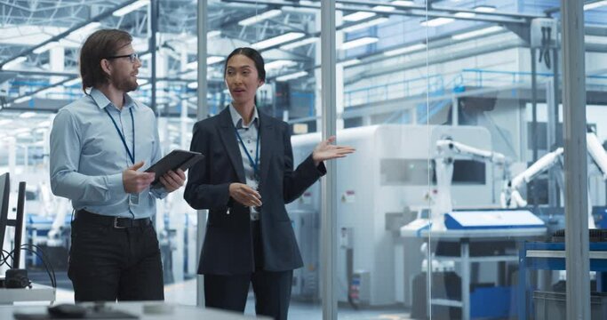 Two Diverse Engineers Walking in Office, Using Tablet Computer at an Electronics Factory. Female and Male Specialists Chatting, Monitoring Robots on a Modern Assembly Line