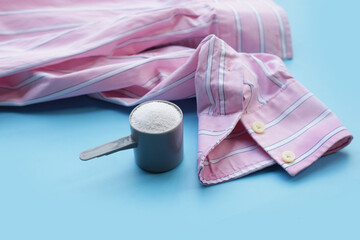 Detergent powder in measuring spoon with shirt on blue background. Laundry concept.