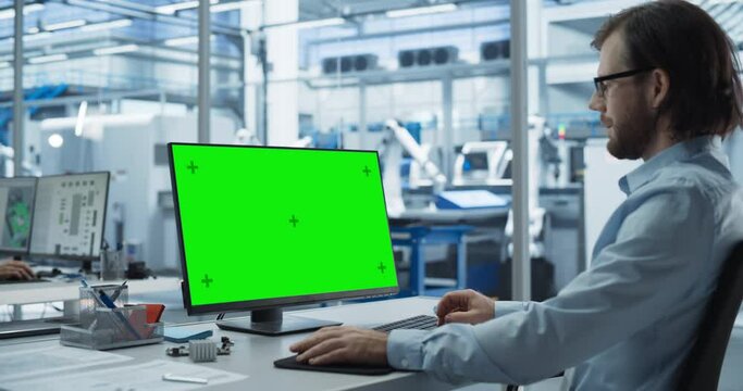 Robotics Engineer Working on a Desktop Computer with Green Screen Chromakey Mock Up Display in a Factory Office. Technological Research and Development Facility with Autonomous Assembly Conveyor