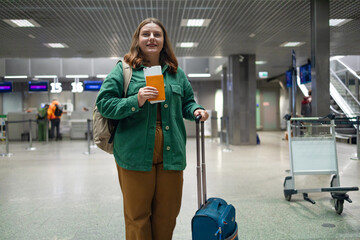 Smiling woman walking with luggage in airport terminal, cheerful female holding passport with...