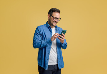 Smiling handsome entrepreneur scrolling social media over mobile phone against yellow background - obrazy, fototapety, plakaty