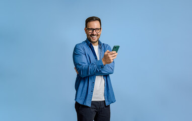 Young businessman texting on smart phone and looking at camera while standing on blue background