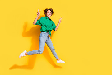 Fototapeta na wymiar Full length photo of funky shiny girl dressed green shirt jumping high showing v-signs empty space isolated yellow color background