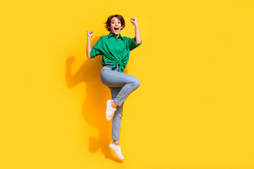 Full length photo of carefree lucky girl dressed green shirt rising fists jumping high empty space isolated yellow color background