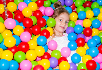 Fototapeta na wymiar Happy girl playing in ball pit on birthday party in kids amusement park and indoor play center. Child playing with colorful balls in playground ball pool.